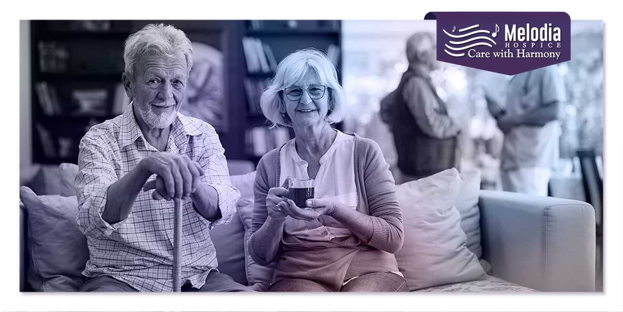 Elder couple on sofa, man with support stick, woman enjoying tea during continuous home care in hospice.